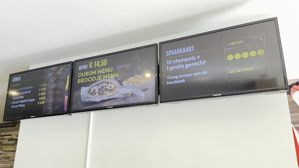 Drei Bildschirme an der Wand im Lokal von Daily Bites in Voeren (Belgien): Döner mit Preisen, Menü für 14,50 € und die Stempelkarte