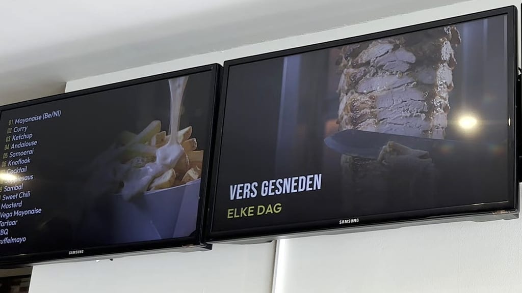 Zwei Bildschirme im Lokal von Daily Bites in Voeren (Belgien): links die Liste der Saucen mit Pommes, rechts «Vers gesneden – elke dag» mit einem Dönerspieß