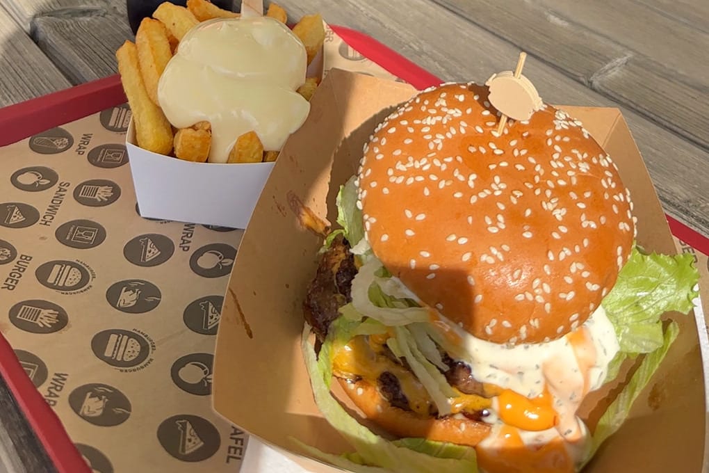 Smash Burger in der Pappschale und Pommes mit Soße auf einem roten Tablett, auf einem Holztisch der Terrasse von Daily Bites in Voeren (Belgien)
