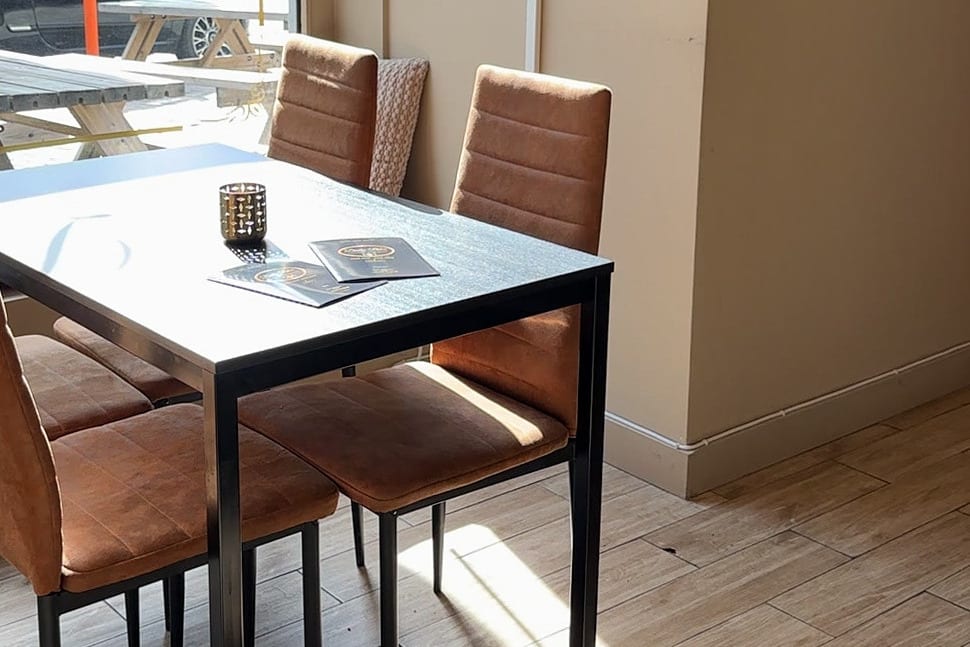 Gastraum von Daily Bites in Voeren (Belgien) im Sonnenlicht: schwarzer Tisch mit zwei Speisekarten und einem Teelichthalter, braune Stühle, vor dem Fenster die Terrasse