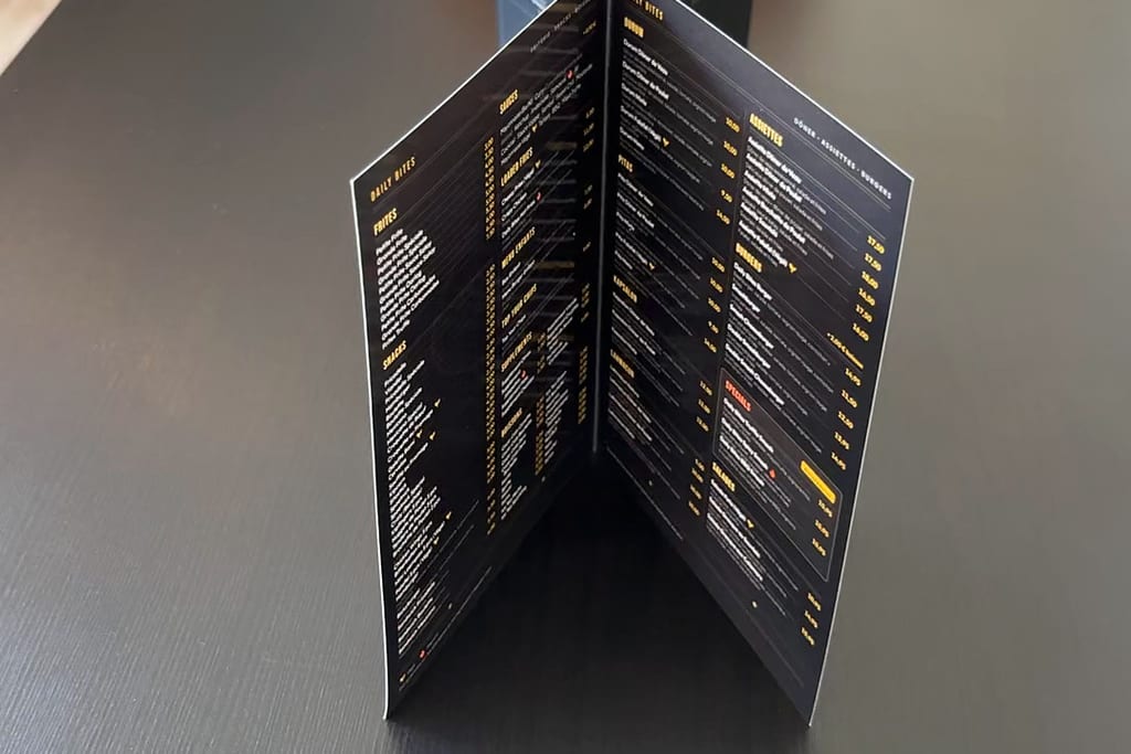 Aufgeklappte Speisekarte von Daily Bites, aufgestellt auf einem schwarzen Tisch, die Innenseiten mit Gerichten und Preisen