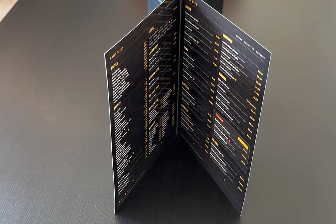 Gedruckte Speisekarte von Daily Bites in Voeren (Belgien), aufgeklappt auf einem schwarzen Tisch, die Innenseiten mit Gerichten und Preisen