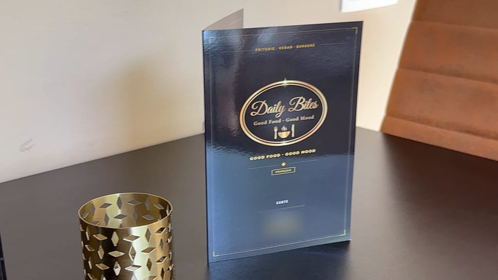 Aufgestellte Speisekarte von Daily Bites in Voeren (Belgien) auf einem schwarzen Tisch, auf dem Cover das goldene Logo, daneben ein goldener Teelichthalter; Adresse und Telefonnummer sind unkenntlich gemacht
