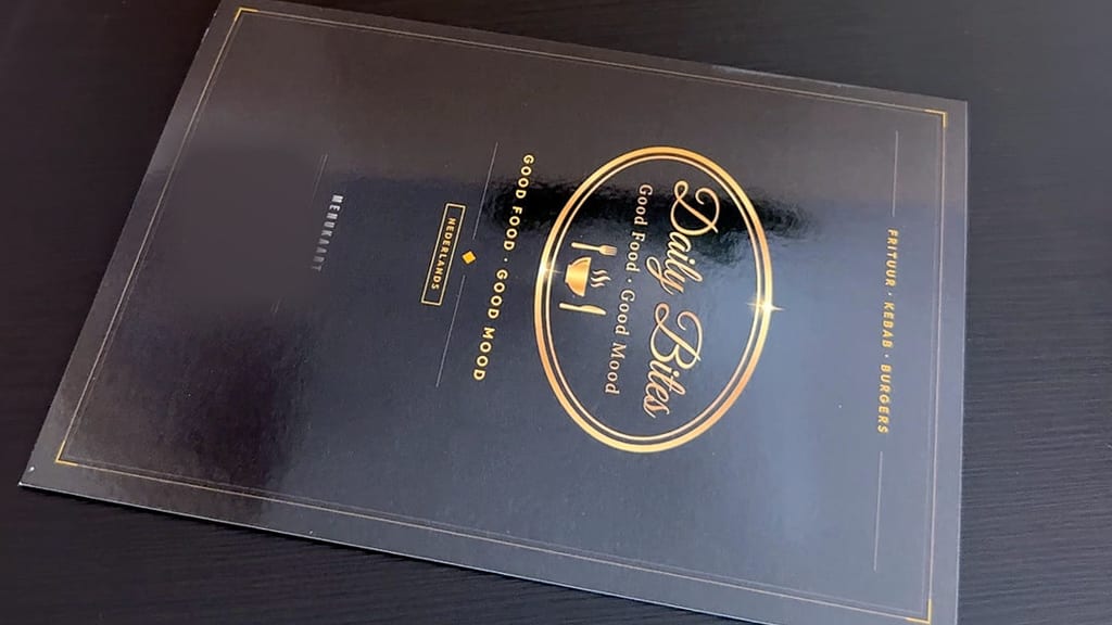 Speisekarte von Daily Bites auf einem dunklen Tisch: das ovale Logo in Gold mit «Good Food · Good Mood» auf schwarzem Cover mit Goldrahmen