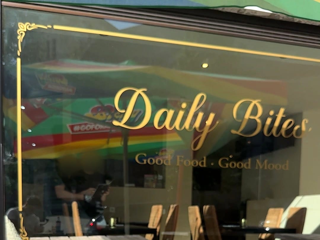 Schaufenster von Daily Bites mit dem Schriftzug «Daily Bites – Good Food · Good Mood» und einem Zierrahmen in Goldfolie, im Glas spiegelt sich die Terrasse