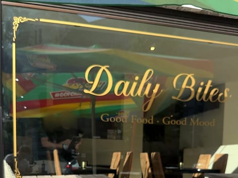 Schaufenster von Daily Bites mit dem Schriftzug «Daily Bites – Good Food · Good Mood» und einem Zierrahmen in Goldfolie, im Glas spiegelt sich die Terrasse