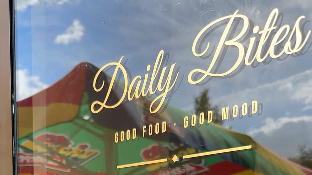 Glastür von Daily Bites in Voeren (Belgien) mit dem Schriftzug «Daily Bites» und «Good Food · Good Mood» in goldener Folie, im Glas spiegeln sich ein Sonnenschirm und der Himmel